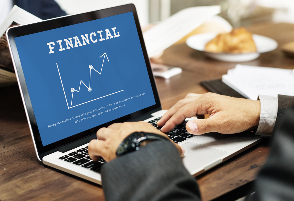Person using laptop showing ‘FINANCIAL’ and rising graph, seated at desk with documents and food in background.