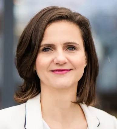 Antje Langsch, with short brown hair and light skin, smiles in a white blazer against a blurred background.