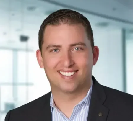 Eric Llorey with short brown hair in a business suit smiles in a modern office with large windows behind him.