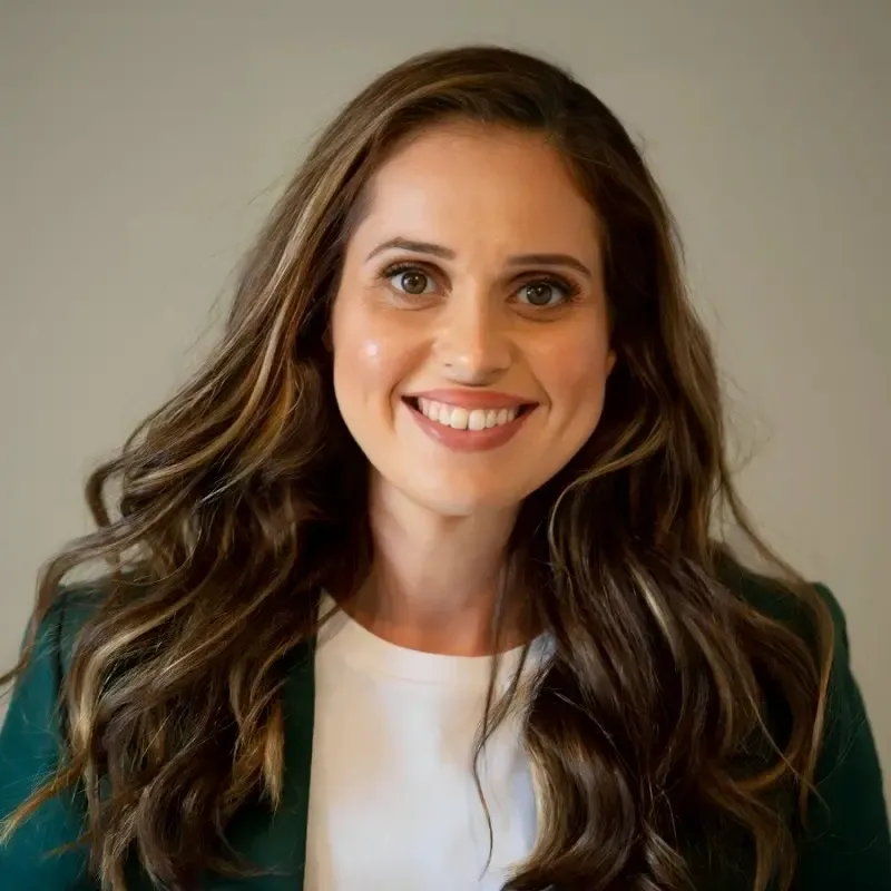 Kylie Kapeleris with long brown hair, in a green blazer and white top, smiling at the camera against a plain backdrop.