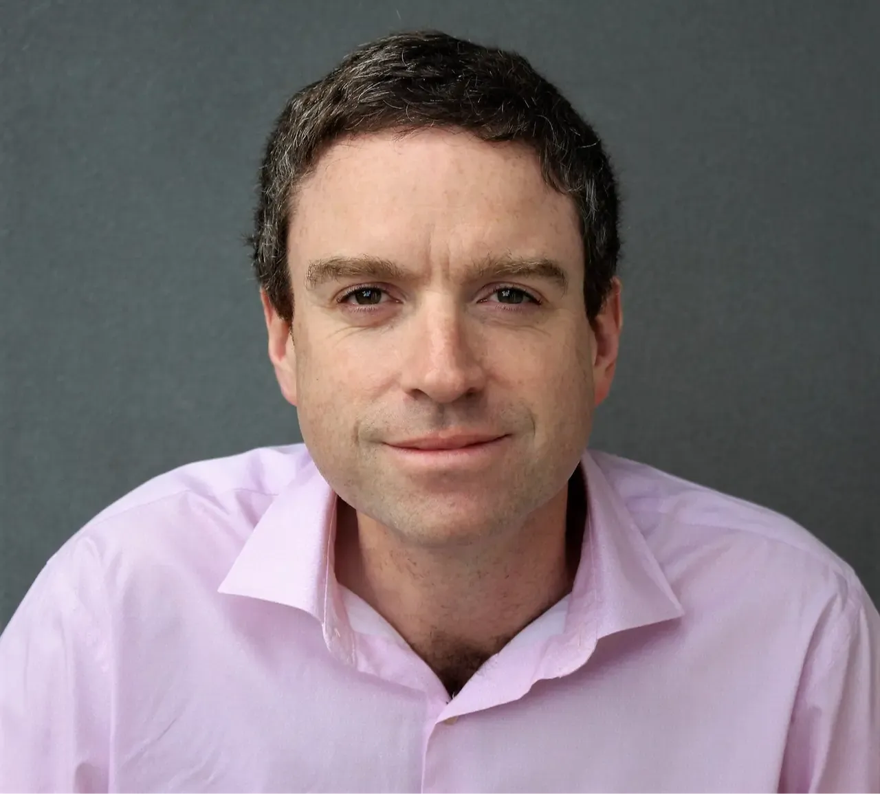 Robbie Jordan, with short dark hair and a light pink shirt, sits against a gray background looking at the camera.