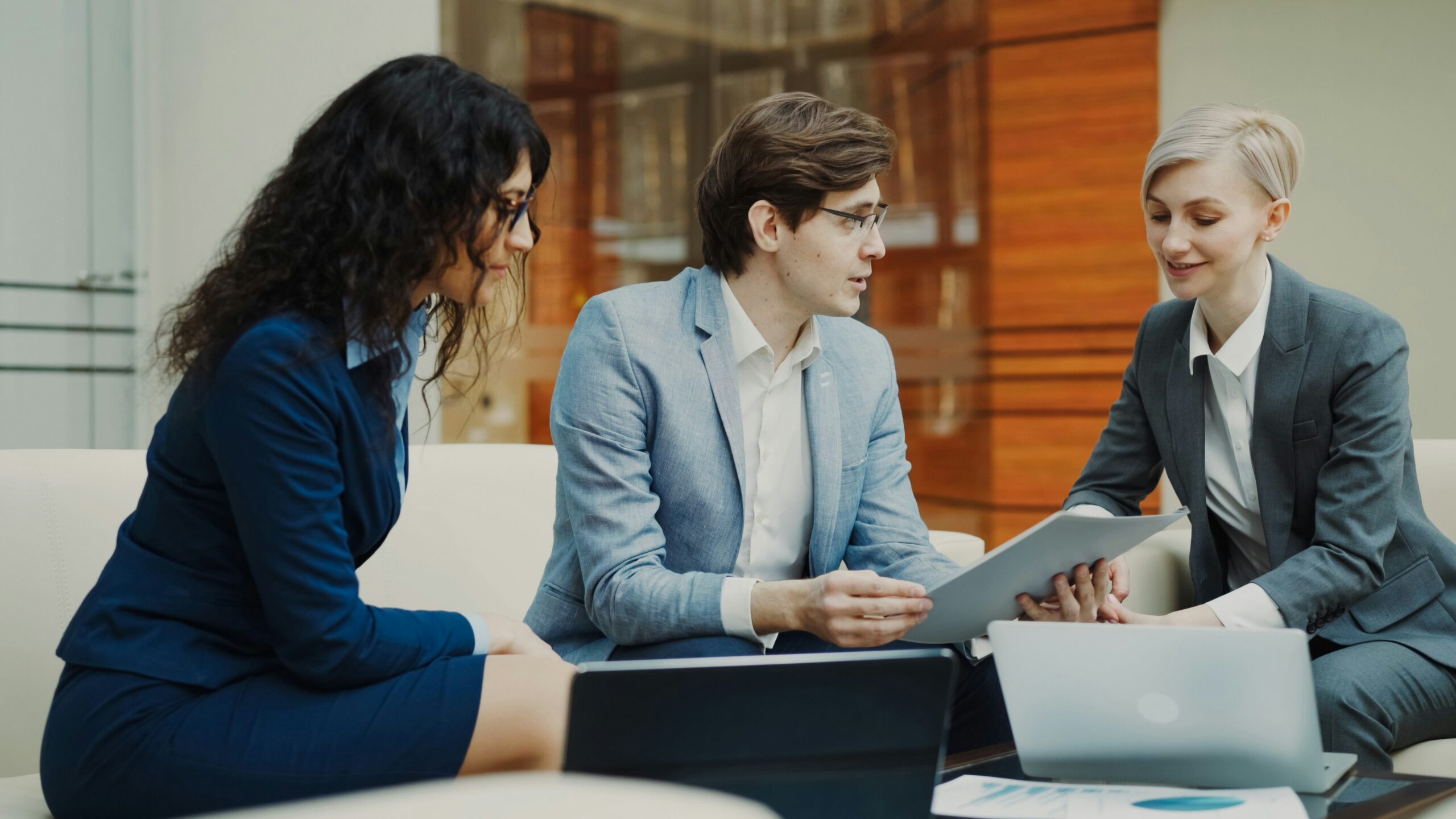 Three people in business attire discussing documents and showcasing finance presentation skills with a tablet and laptop.