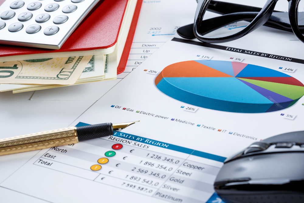 A desk with financial documents, pie chart, calculator, pen, cash, glasses & mouse illustrating EBITDA sales data.
